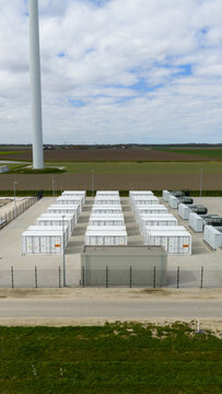 Dronten, Netherlands - 04 April 2026: Aerial view of the Olsterdwarspad battery energy storage system with rows of white containers and a wind turbine mast under a cloudy sky.