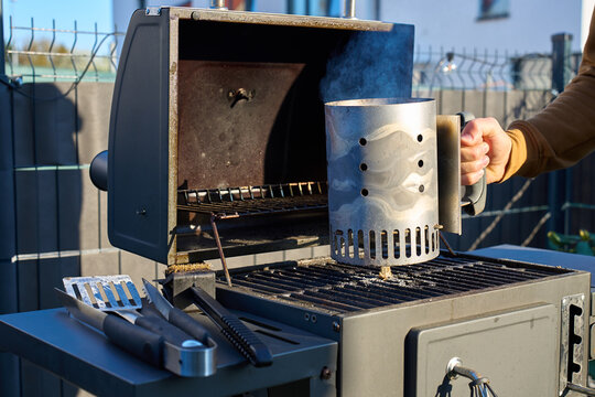 Person holds smoking charcoal chimney on grill with barbecue tools on side shelf
