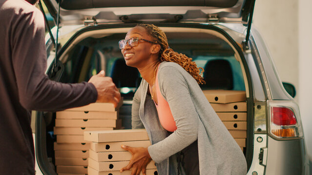 Female courier delivering pizza boxes stack from car trunk, taking huge pizzeria order in piles out of vehicle. Takeaway delivery worker giving packages to customers standing. Handheld shot.