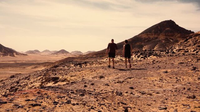 Panorama of Egypt&rsquo;s Black Desert with basalt formations and two walking tourists. Wide panoramic shot of Egypt&rsquo;s Black Desert, showing numerous dark basalt formations and scattered rocks 