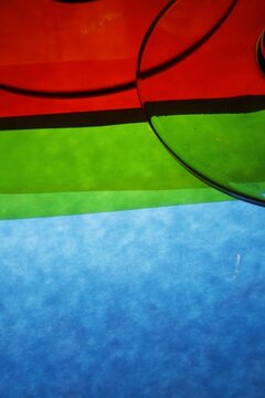 Primer plano abstracto de las bases de copas de vidrio donde la luz atraviesa el cristal y proyecta intensos tonos rojo, verde y azul sobre un fondo de franjas horizontales geom&eacute;tricas.