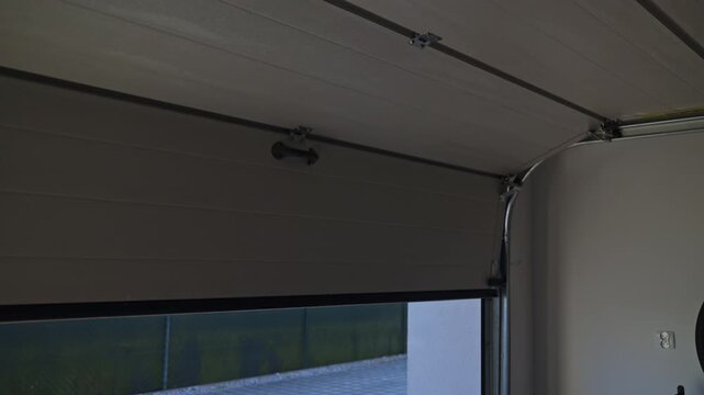 Sectional garage door is closing from inside with yard, fence and trees visible outside. Concept of residential garage access and property security