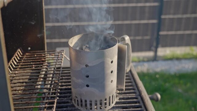 Hand shakes chimney starter over outdoor grill with smoke, sparks and charcoal. Barbecue preparation and outdoor cooking concept