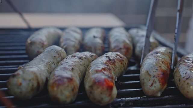 Metal tongs turn sausages on a barbecue grill. Grilling, food preparation and outdoor cooking concept