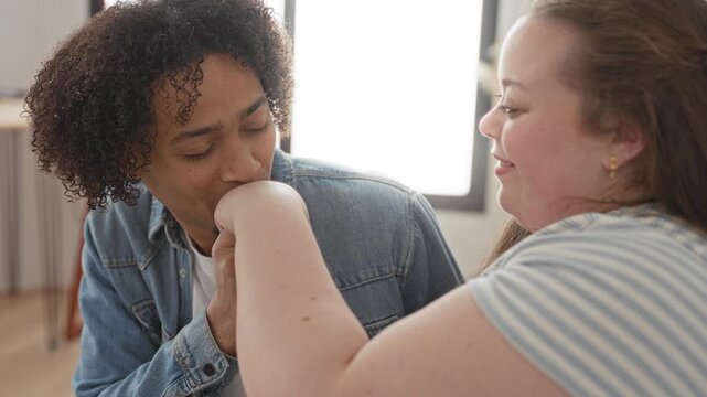 Man kissing plus size woman's hand in a sunlit studio, visible hand holding gesture and denim jacket; tenderness connection.
