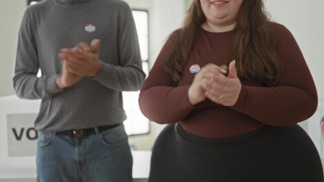 Man and woman clapping with 'i voted' stickers beside a ballot box and vote sign in a polling station interior inside a building; pride civic duty.