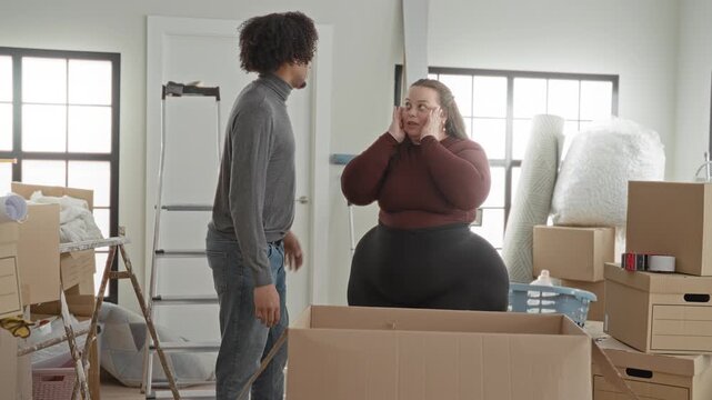 Man and plus size woman stand by open moving box, man hand on forehead and woman touches face in building; moving day frustration together.