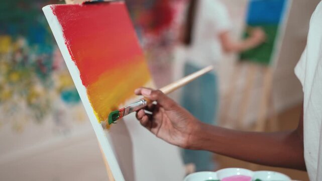 Closeup of african boy hand paint canvas with warm tone color while caucasian girl in casual outfit drawing cool tone colors behind at colorful stained wall. Creative activity concept. Edification