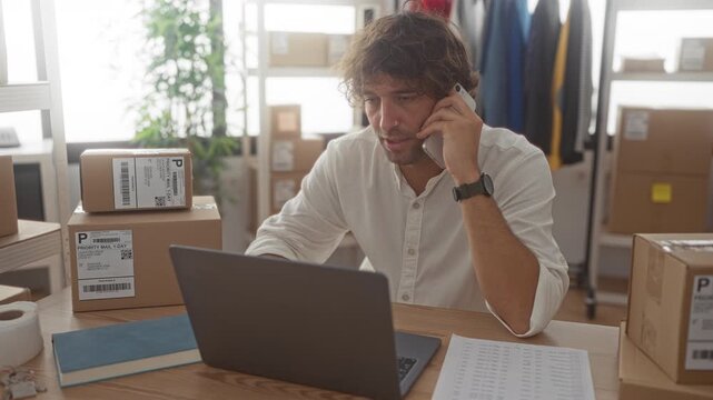 Man holds phone to ear with hand and types at laptop amid stacked parcels and shipping boxes in studio; focus.