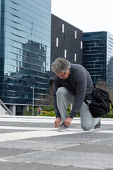 Senior man kneeling tying sneaker lace on patterned plaza near glass towers with black shoulder bag © wavebreak3