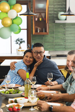 Diverse friends sharing celebratory meal at home on wooden dining table with wine and balloons