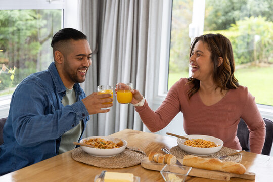 Couple clinking orange juice glasses while sitting at wooden dining table with pasta and baguette