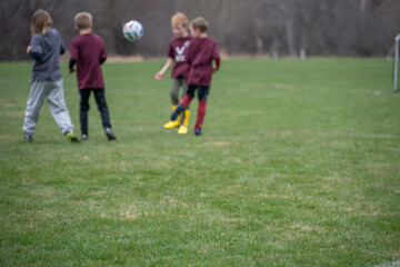 Fototapeta premium Kids play soccer on a grassy field during a cloudy day in a local park with other players in the background