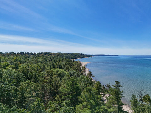 Aerial view of Maple Bay Natural Area with dense green forest meeting the sandy beach and blue water of Lake Michigan at Williamsburg, Michigan, United States.