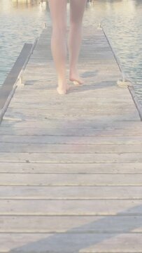 Vertical video: Teen girl walking along dock, bare feet, first step starting travel shot at sunset