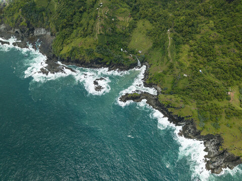 Aerial view of Banyutibo Water Fall cascading over dark rocks into the turquoise ocean along a lush green coastline with terraced fields Girisubo, Special Region of Yogyakarta, Indonesia.