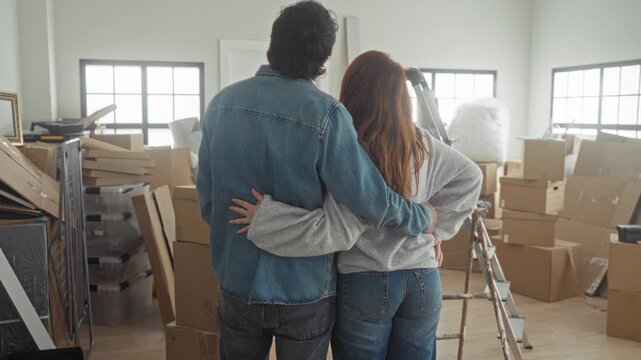 Man and woman stand amid stacked cardboard boxes and a ladder in a building, man holds woman with arm around her waist while she flips hair and smiles at camera; new home joy.