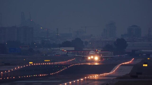 VALENCIA, SPAIN - JANUARY 23, 2026: Head-on shot of a passenger airplane with bright lights landing on a wet runway at dusk. The aircraft approaches, touches down, and rolls towards the camera in a