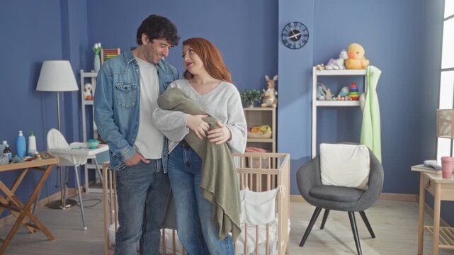 Woman kissing partner while cradling a swaddled newborn by a wooden crib in a nursery building with toys, changing table and armchair; tenderness bonding warmth.