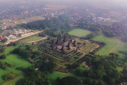 Aerial view of Prambanan Temple complex with its stone spires surrounded by lush green lawns and tropical trees under a hazy morning sky in Kecamatan Prambanan, Daerah Istimewa Yogyakarta, Indonesia.