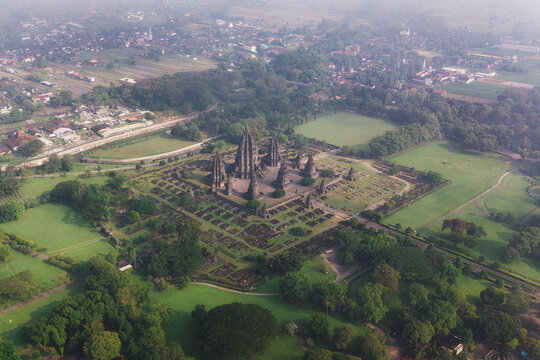 Aerial view of Prambanan Temple featuring ancient stone towers, lush green parkland, and surrounding village under a soft morning haze in Kecamatan Prambanan, Daerah Istimewa Yogya, Indonesia.