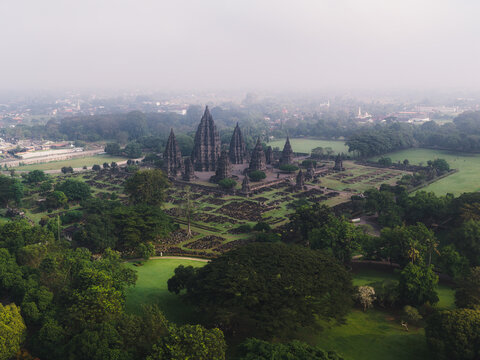 Aerial view of the Prambanan Temple complex featuring ancient stone spires and lush green parkland under a misty sky at Kecamatan Prambanan, Daerah Istimewa Yogya, Indonesia.