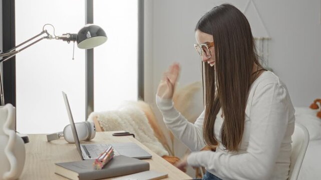 Woman student waves hands over laptop while smiling and wearing glasses at a bedroom desk in building; focused study.