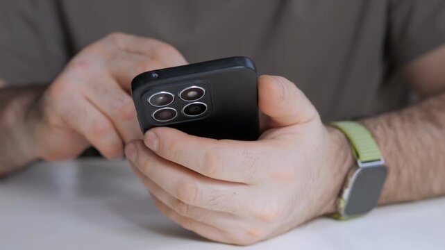 Hands hold black smartphone with triple camera. Watch with green strap on wrist. Person taps screen while seated at table. Focus on device use and interaction. No animals or plants visible