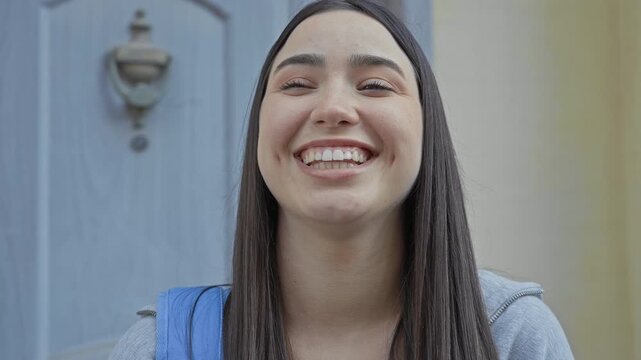 Woman hand covering face in a building doorway, smiling and laughing with visible nails, ring and backpack strap; shyness joy.