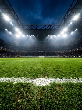 Professional football stadium at night from a low perspective behind the goal line showing bright floodlights, crowded spectator stands, and green grass turf under a dark sky.