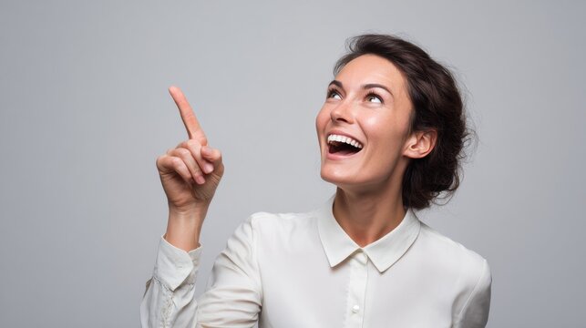 Smiling woman in white shirt pointing upwards against gray background