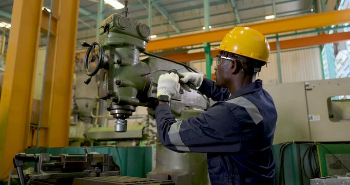 African adult male technician crouches beside large milling machine, aiming air blow gun toward work area during maintenance inspection, equipment cleaning and safe production setup in slow motion
