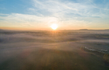 Fototapeta premium Aerial view captures bright sun cresting a misty valley landscape, casting warm light across green fields and distant mountains at dawn