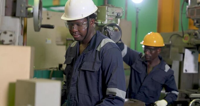 African adult male operator studies machine condition at industrial workstation as coworker reaches toward equipment behind, factory activity, PPE discipline, process monitoring, output support.