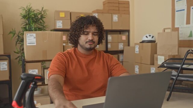 Man at laptop holding papers in building amid stacked cardboard boxes and barcode scanner, hand turning a sheet; order management concern.