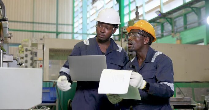 Two African adult male engineers study laptop and printed sheet near production machine, coordinating inspection result, technical review and data driven manufacturing decision inside