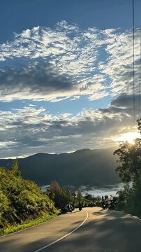 A winding road leads to distant mountains beneath a sky filled with textured clouds and the glow of the rising or setting sun.