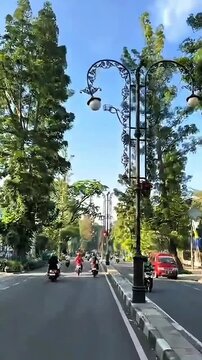 A sunlit street lined with trees and ornate lampposts leads to a distant archway, creating a peaceful urban scene under a bright blue sky.