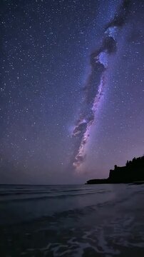 The night sky explodes with stars above a tranquil ocean, reflecting the Milky Way's ethereal glow onto the dark, silhouetted coastline in this captivating vista.
