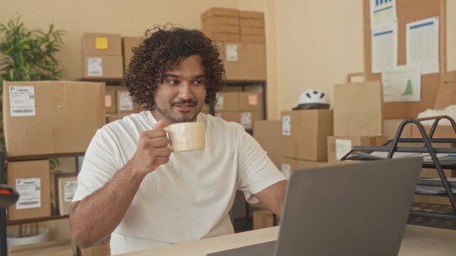 Man holding coffee mug and typing on laptop among stacked cardboard boxes in building; entrepreneurial calm.
