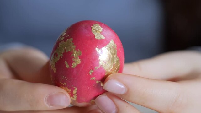 Close-up of hands holding a red egg with gold patterns. Finger gently touches the egg's surface. Soft lighting highlights the egg's texture and color. Background is blurred, focusing on the egg