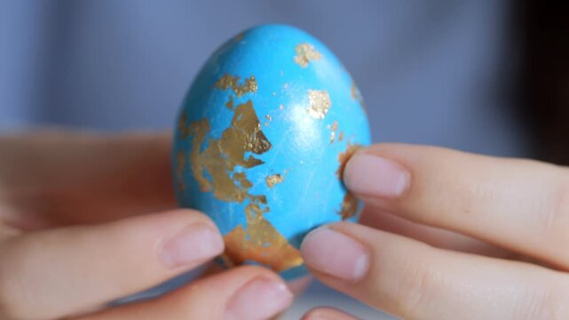 Hands gently hold a blue egg with gold leaf patterns. Soft focus background emphasizes the egg's ornate surface. Fingertips cradle the egg, showing delicate care