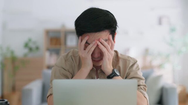 Young Asian man tired bored sad upset fatigued working on laptop in living room at home