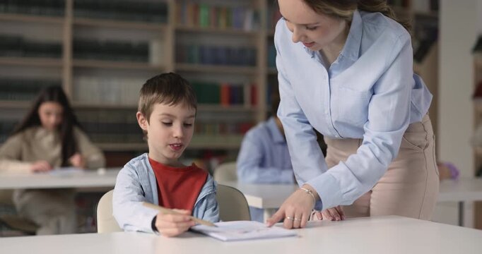 Female teacher assisting with classwork to schoolboy child making schoolwork during lesson in classroom. Education in primary school, elementary learning, early childhood skill development concept