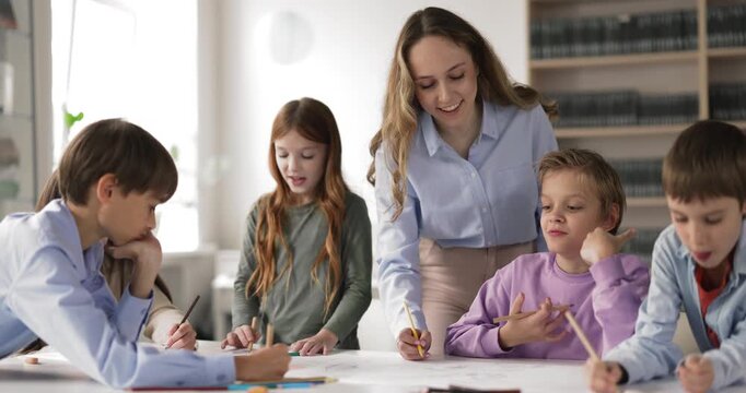 Female teacher helps to diverse children drawing with pencils on paper in classroom. Learning process, skill development, creativity, educational support for primary school or art class pupils concept