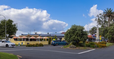 Obraz premium Street scene in Raglan, New Zealand, featuring a white Peugeot car, a blue Holden car, a restaurant with tiki umbrellas, and a war memorial on a sunny day.