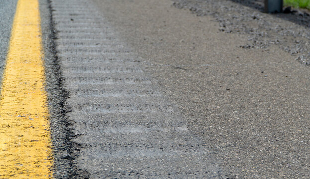 Detailed close-up of a textured asphalt road surface with a bright yellow lane marking and a subtle rumble strip, illustrating infrastructure, wear, and travel.