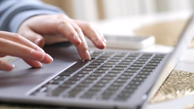 Hands typing on laptop keyboard. Fingers press keys with precision. Soft light glows on silver laptop surface. Background blurred with warm tones. Close-up view of workspace activity