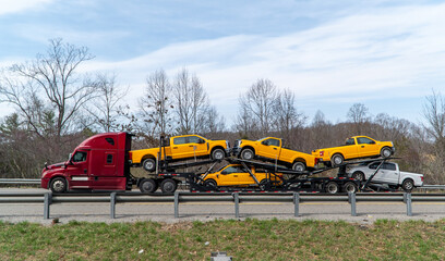 Red semi-truck hauling a multi-level car carrier loaded with several new yellow and white Ford pickup trucks driving on a highway, symbolizing automotive transport and vehicle delivery. © Michael