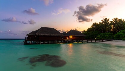 Fototapeta premium Scenic dusk view showcases overwater bungalows illuminated at sunset, adjacent to a tropical beach, with calm ocean and palm trees